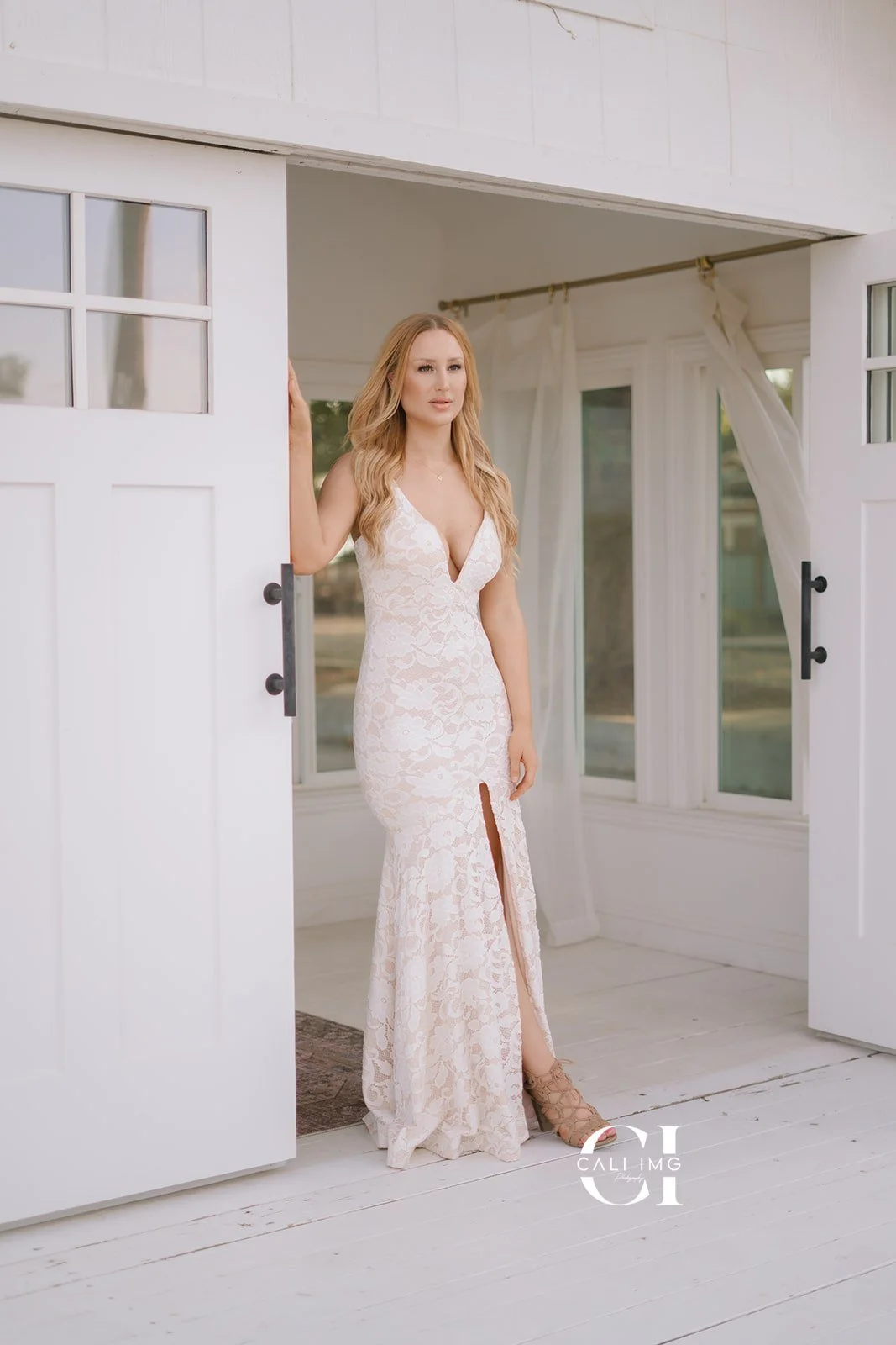 A woman in a white lace dress with a slit standing in a doorway, looking outside in a bright room with white walls and curtains.
