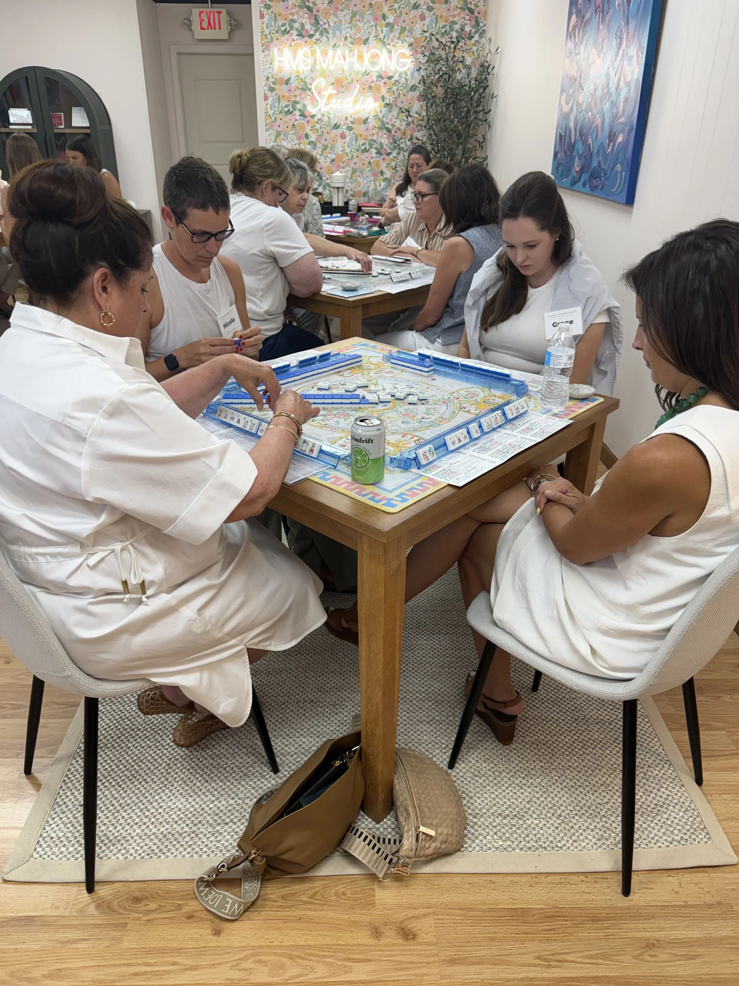 Multiple women playing the game of Monopoly at a wooden table in a decorated room with a floral wallpaper at HMS Mahjong Studio Westmont. There are personal bags and a water bottle on the floor.