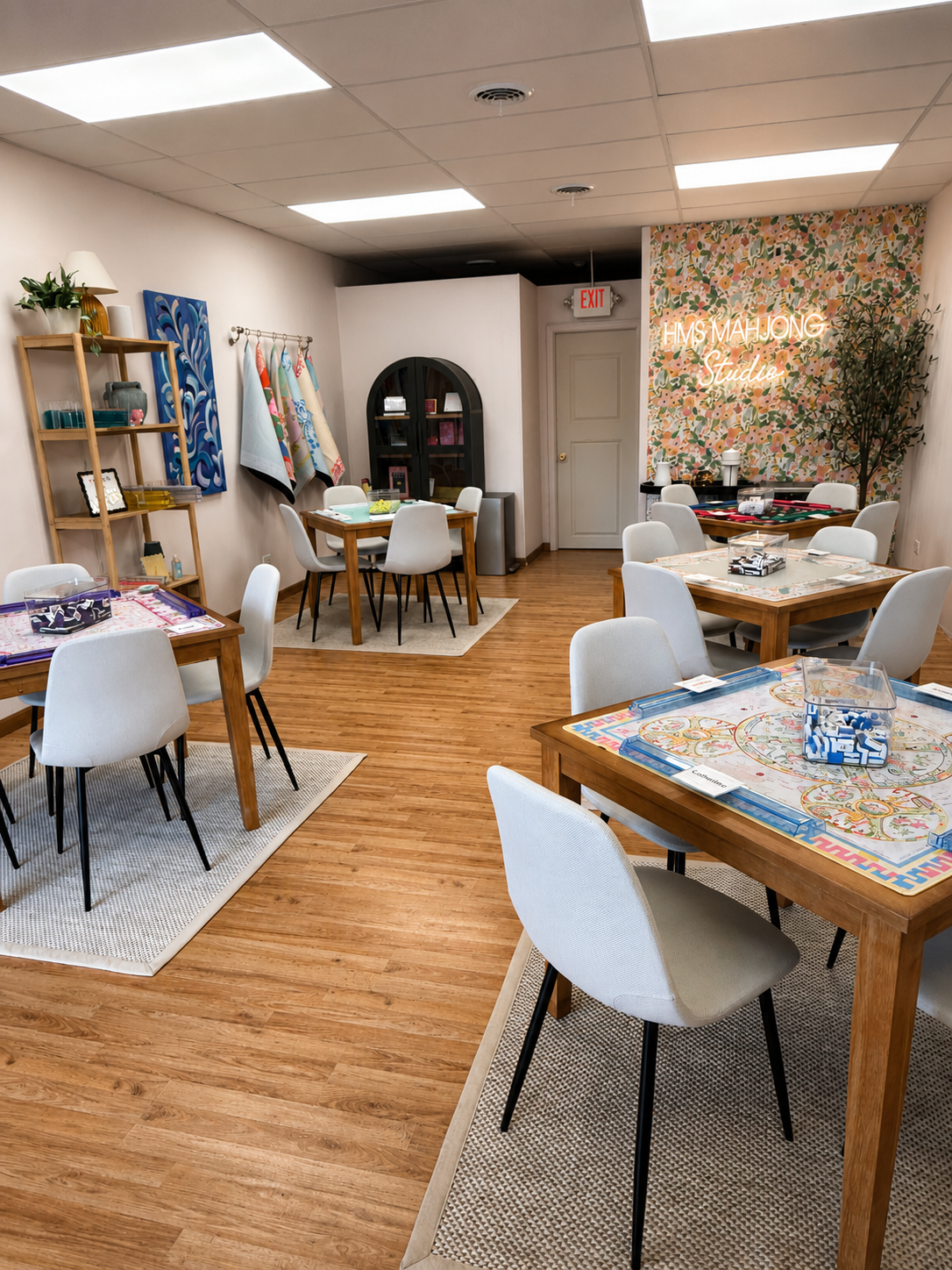 Art studio with tables and chairs, colorful wall art, and a neon sign that reads 'HMS Mahjong Studio.'