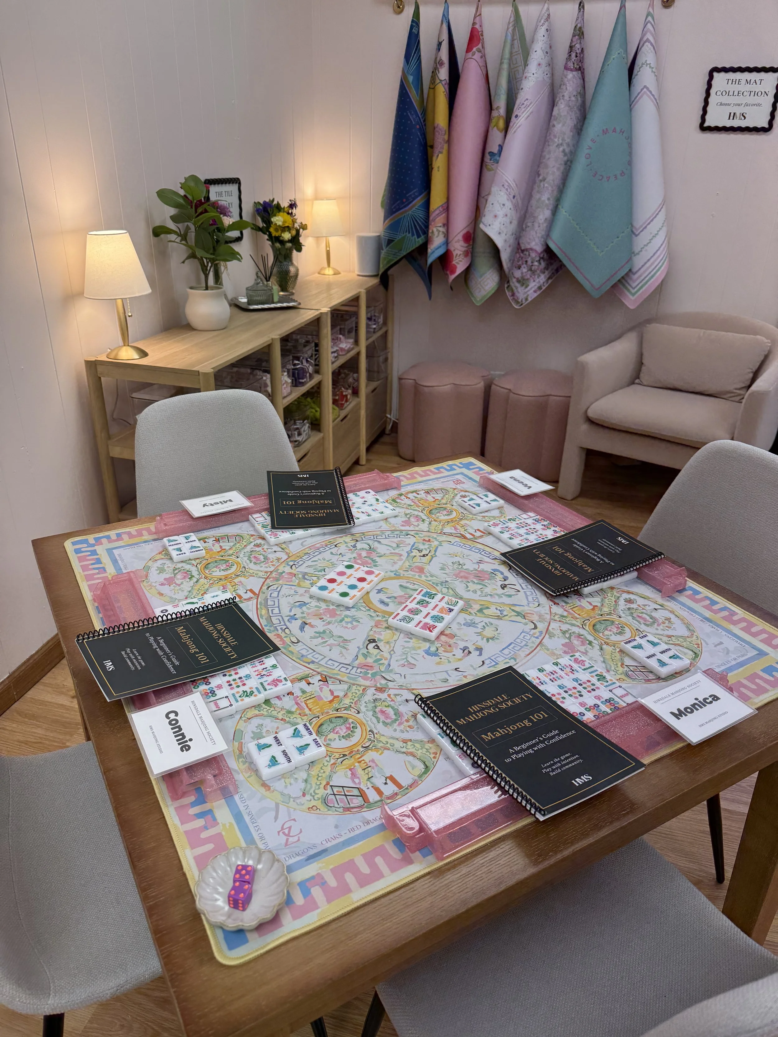 A group of people playing a Mahjong game around a table with colorful Mahjong tiles, rulebooks, and score sheets, in a cozy room with pink walls, decorated with hanging floral and pastel-colored towels, small lamps, and a couch at HMS Mahjong Studio.