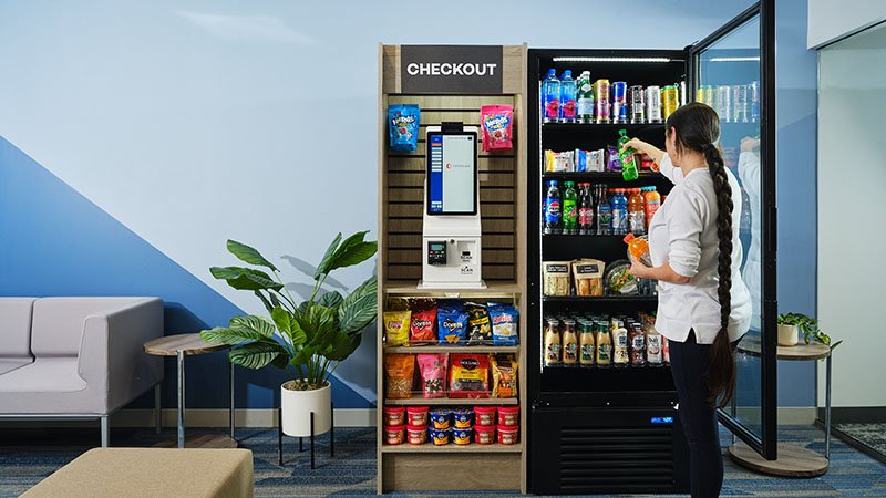 Woman shopping for snacks at a micro market with a checkout kiosk nearby in an office lounge area.