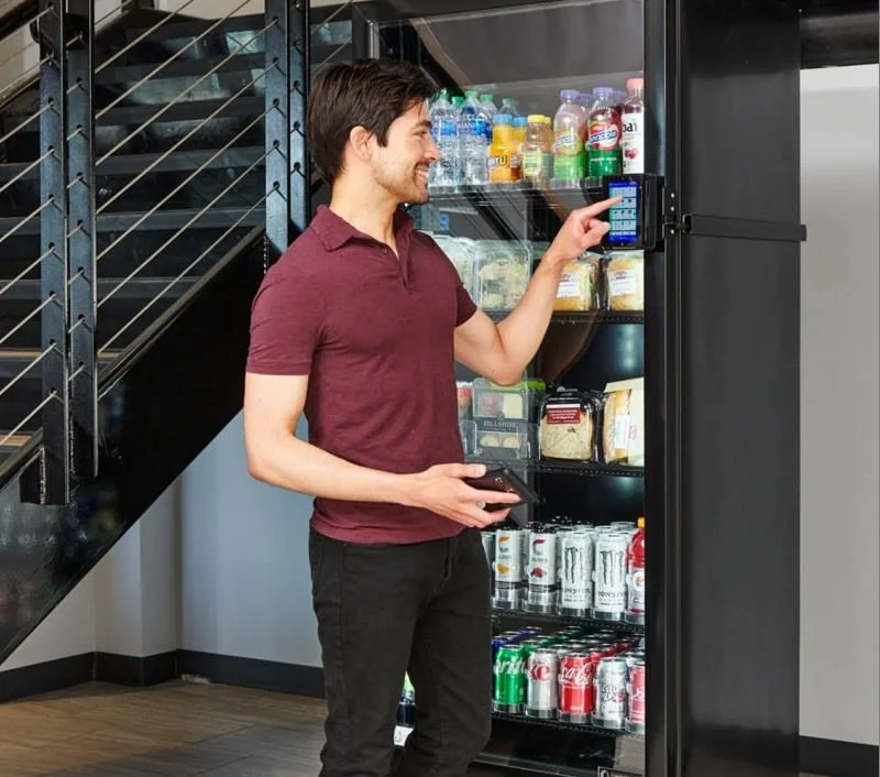 A man in a maroon polo shirt using a touchscreen on a smart cooler stocked with drinks and snacks.