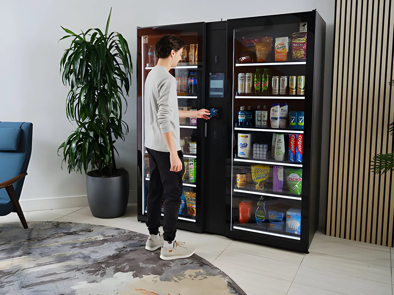 Person using a vending machine for snacks and drinks in an office lounge area. There is a large potted plant to the left and a blue chair partially visible on the left side. The vending machine has snacks and beverages visible inside and is situated 