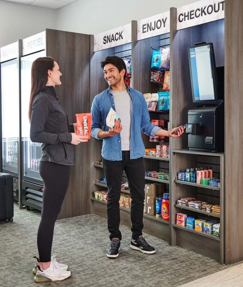A man using a self-checkout kiosk in an airport convenience store, smiling, while a woman in a grey jacket looks at him holding a large snack bar, with shelves of snacks and drinks behind them.