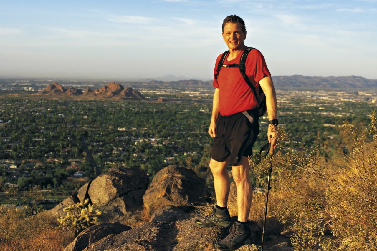A man in hiking gear standing on a rocky hillside overlooking a desert cityscape with mountains in the distance.