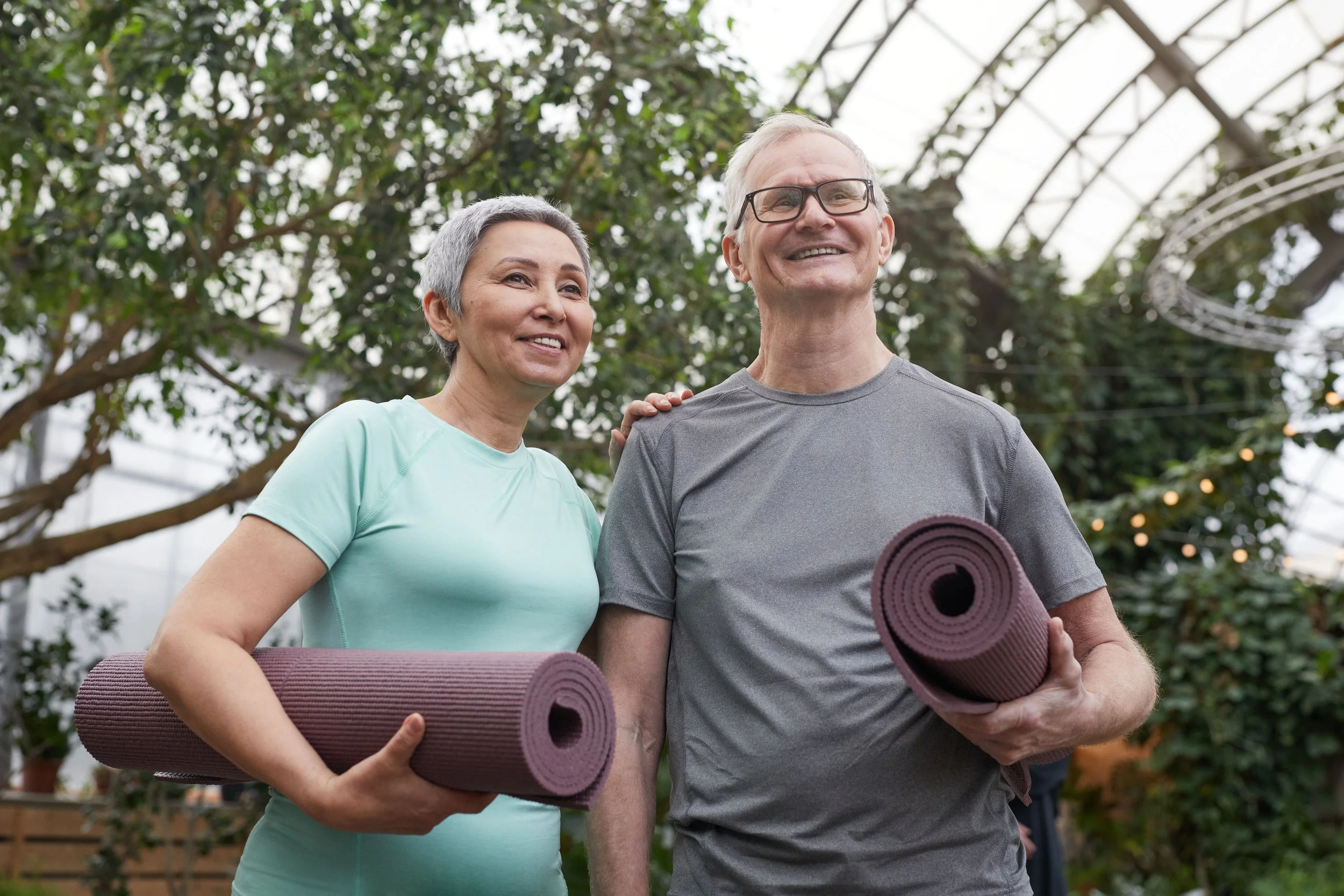 Active older couple holding yoga mats in a garden, representing healthy movement and spine wellness.