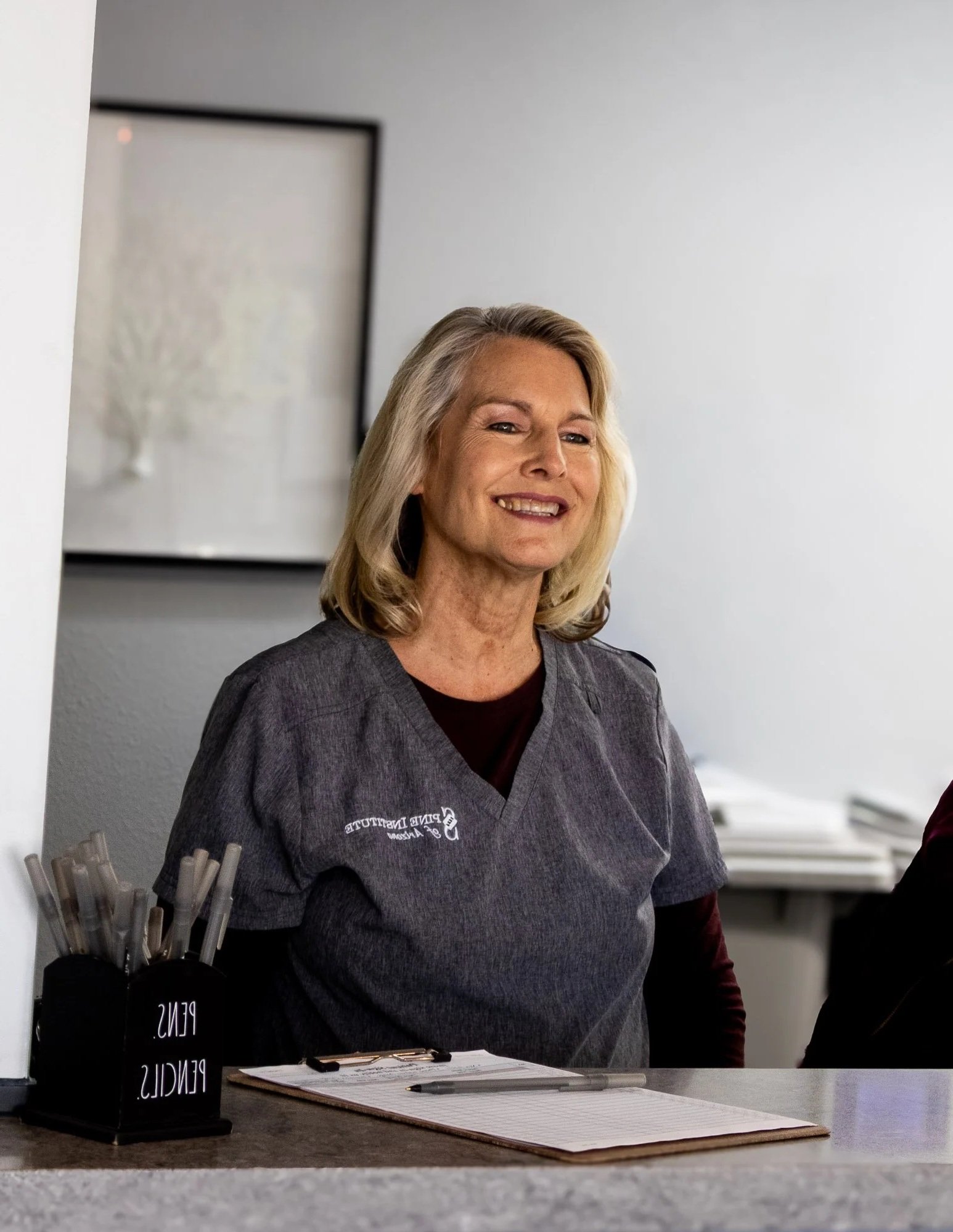 A woman with blonde hair smiling behind a counter with pens, a notepad, and a pen on it. She is wearing a grey scrub top with some white text on it.