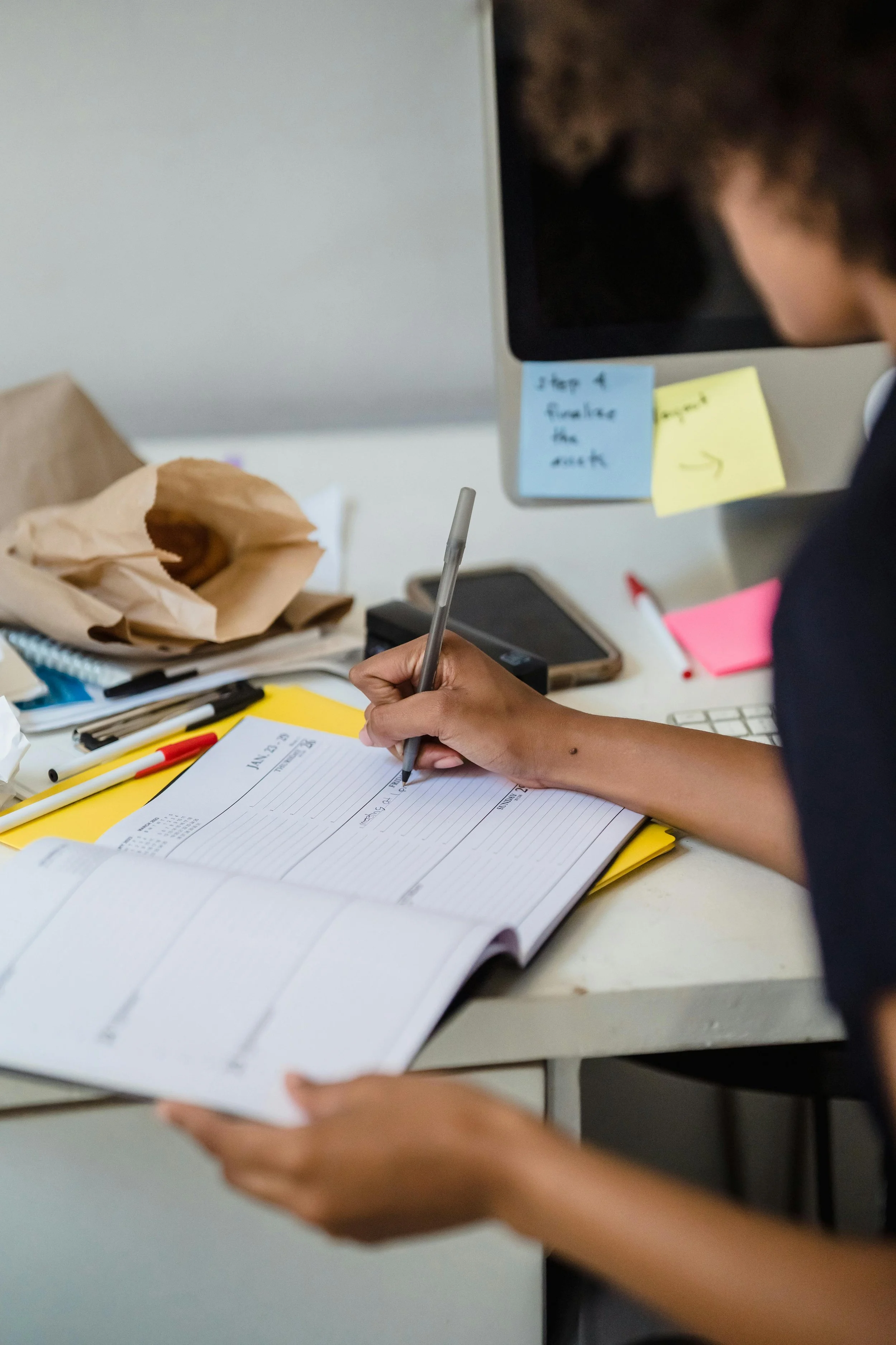 Close-up of a black woman writing in a planner at a desk, focusing on organization in a work environment. Dr. Mary Woodson provides flexible supervision scheduling in NC & CO.