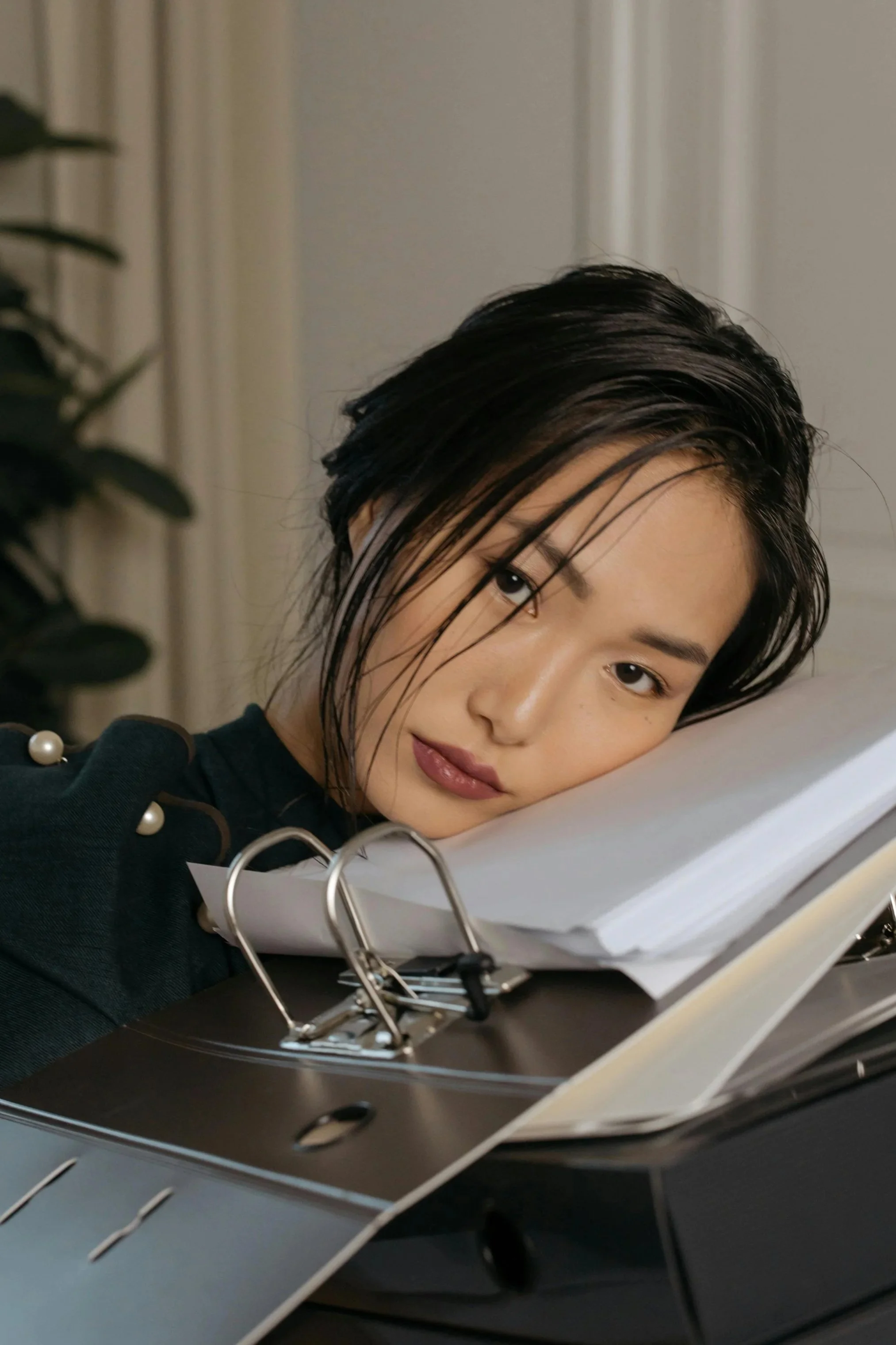 A young woman with wet dark hair and makeup, resting her head on a folder filled with papers on a desk.