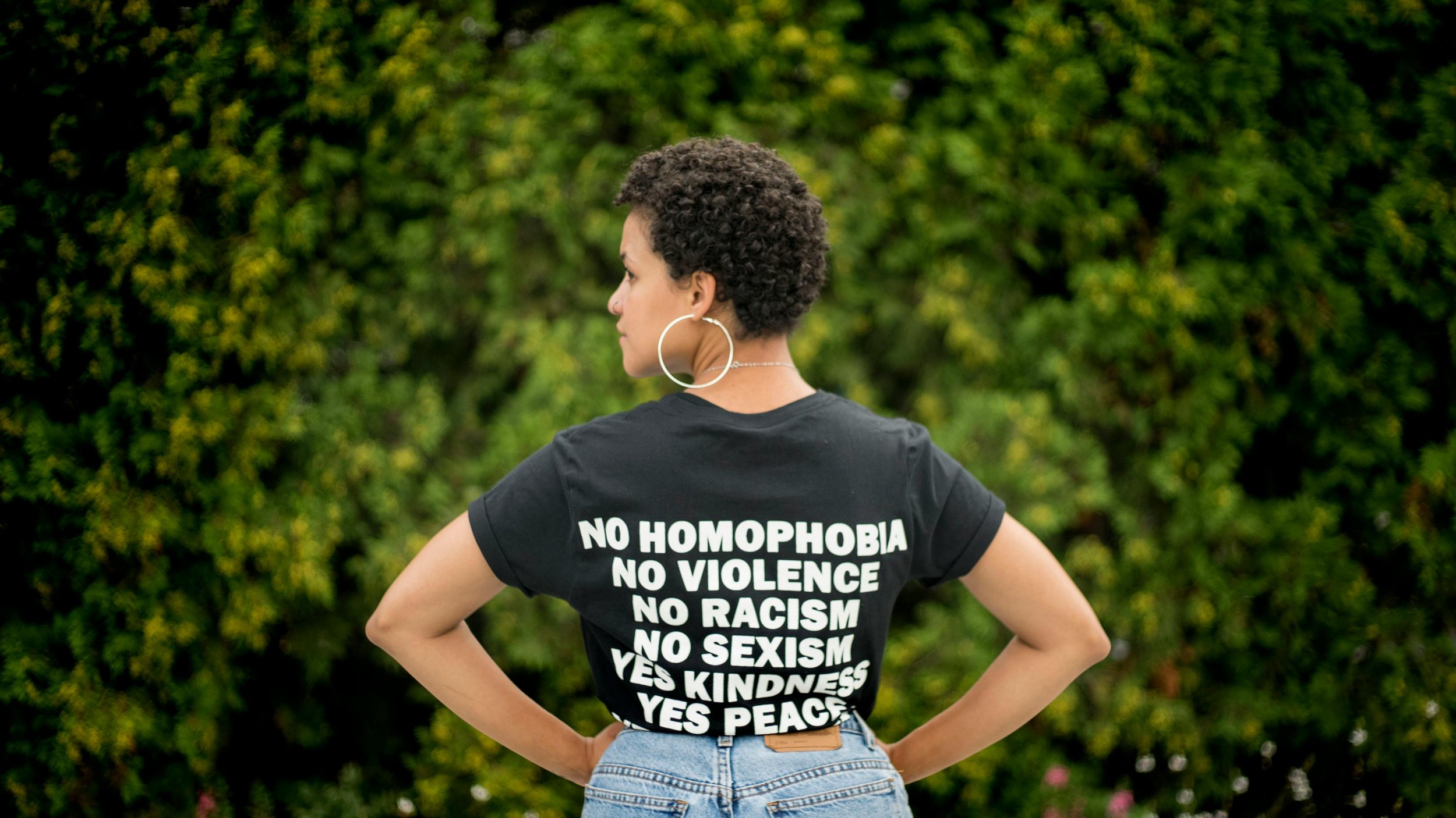 Woman with short curly hair wearing a black T-shirt with message opposing discrimination, standing outdoors with trees in the background. Radical, liberation therapy for LGBTQIA+ & BIPOC with Dr. Mary Woodson, queer black woman therapist.