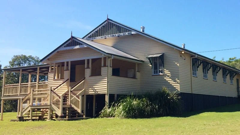 A historic wooden building with a sign indicating it was established in 1908, featuring a staircase and a porch, surrounded by greenery on a clear day.
