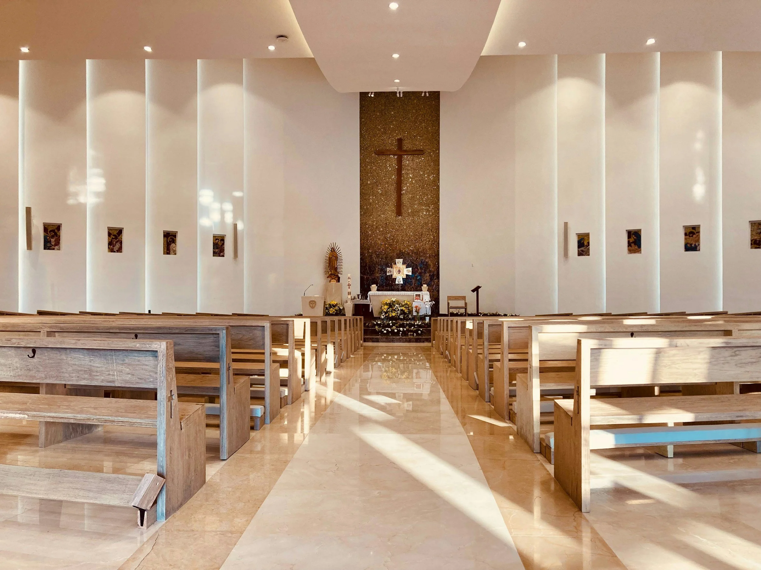 Interior of a church or chapel with wooden pews, a cross on the wall behind an altar, religious icons on either side, and a white and gold cross at the center of the altar.