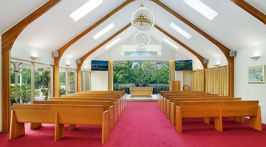 Interior of a chapel with wooden pews, a red carpet, large windows, and modern chandeliers hanging from the ceiling, with a view of trees outside.
