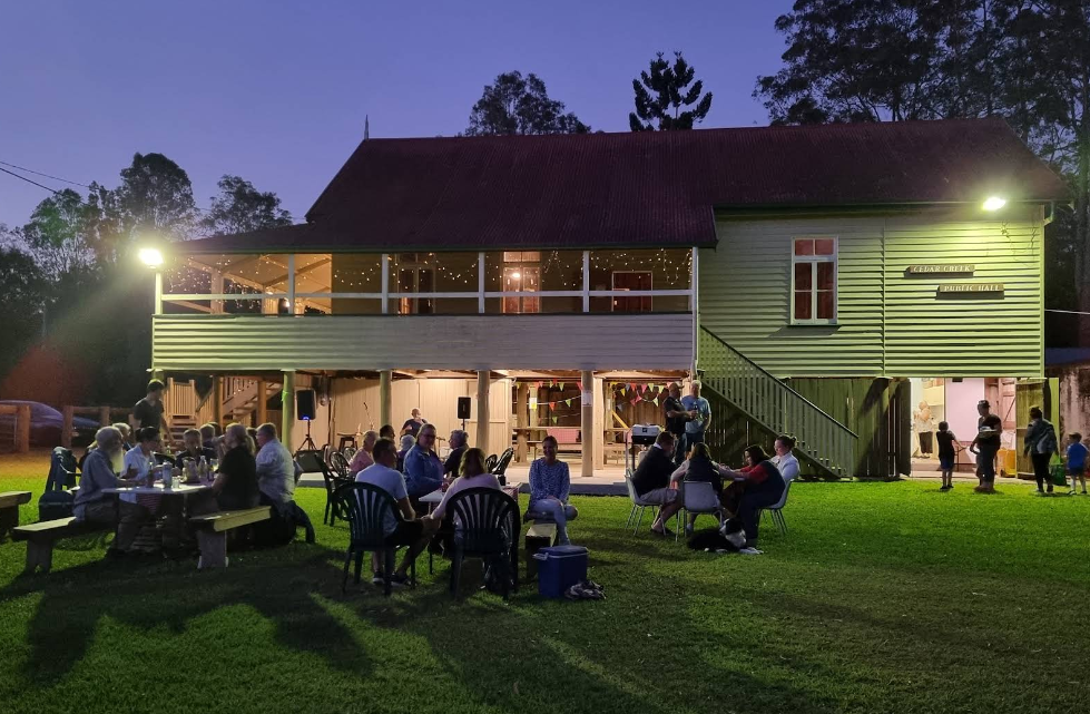 People gathered outdoors at night for a social event, sitting at tables on a grassy area in front of a two-story green building with a balcony, illuminated by lights.