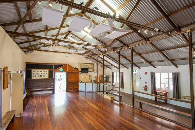 Empty rustic indoor hall with wooden floors, exposed wooden beams, hanging acoustic panels, and large windows with curtains.