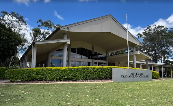 Exterior of the Mt. Grant Crematorium and Chapel building with a large glass facade, surrounded by a well-maintained lawn and trees, under a partly cloudy sky.