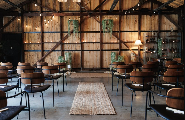 Indoor event space with wooden walls and ceiling, decorated with string lights and hanging greenery. Rows of chairs face a small stage, with a beige rug in the center aisle, and a bookshelf with decorative items and a floor lamp in the corner.