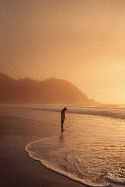 A person stands alone on a beach near the shoreline at sunset, with mountains in the background.