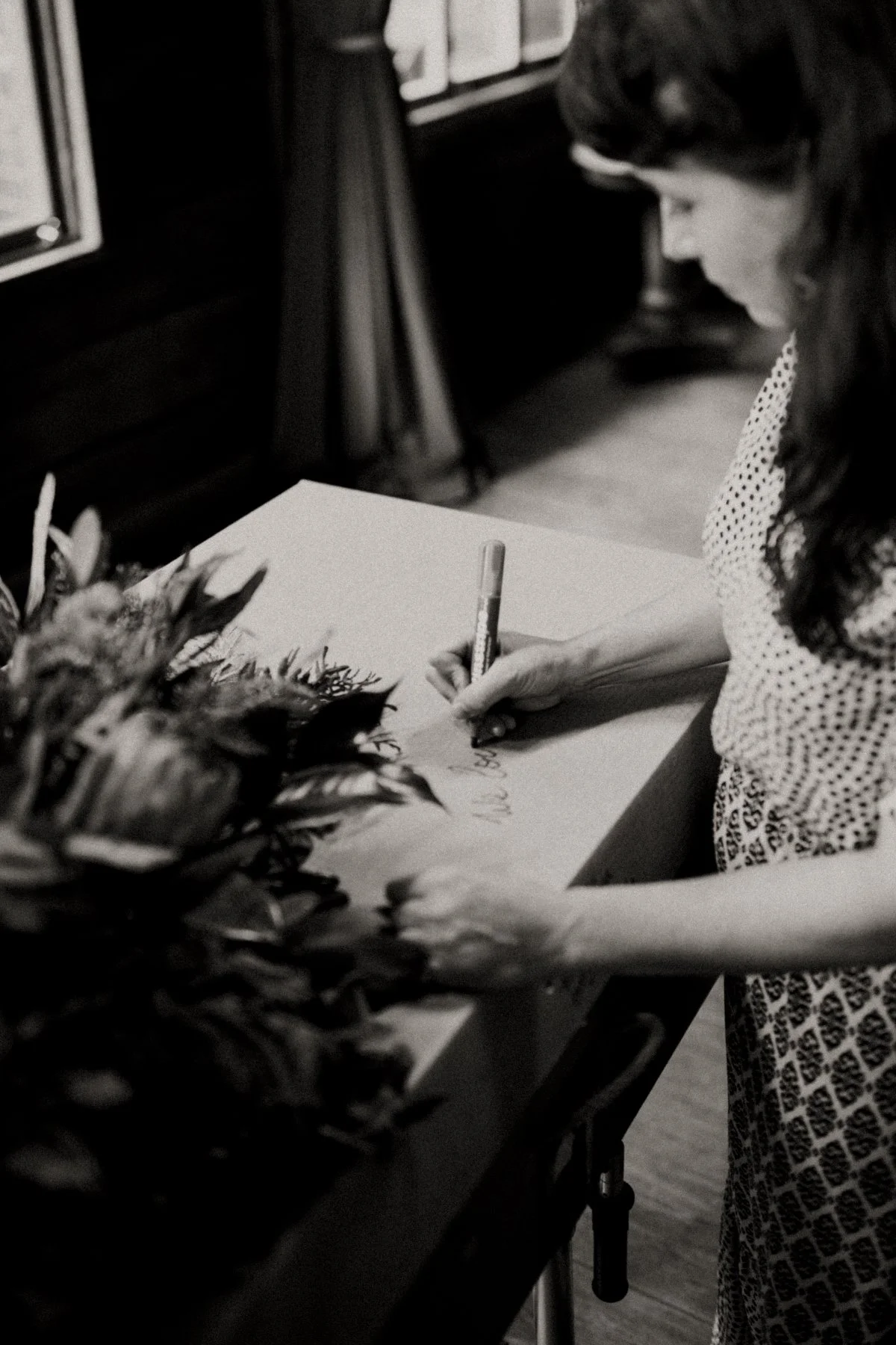 A woman in a polka-dot dress is writing on paper on a table, with a flower arrangement nearby.