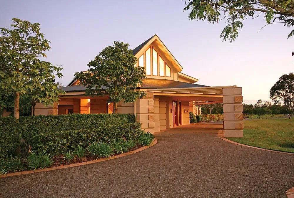A modern house with a triangular window on the roof, surrounded by trees and a well-kept garden, during sunset.