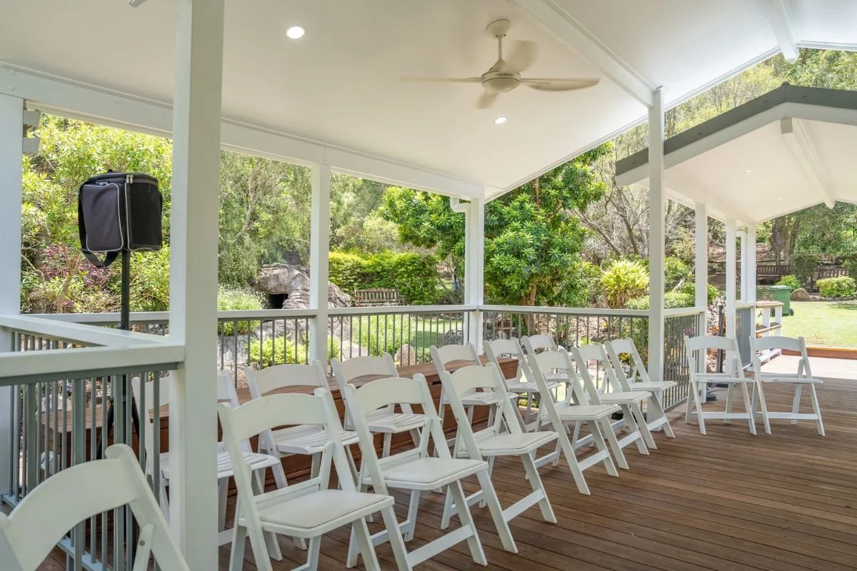 Covered outdoor patio with white wooden chairs arranged in a row, surrounded by greenery and trees.