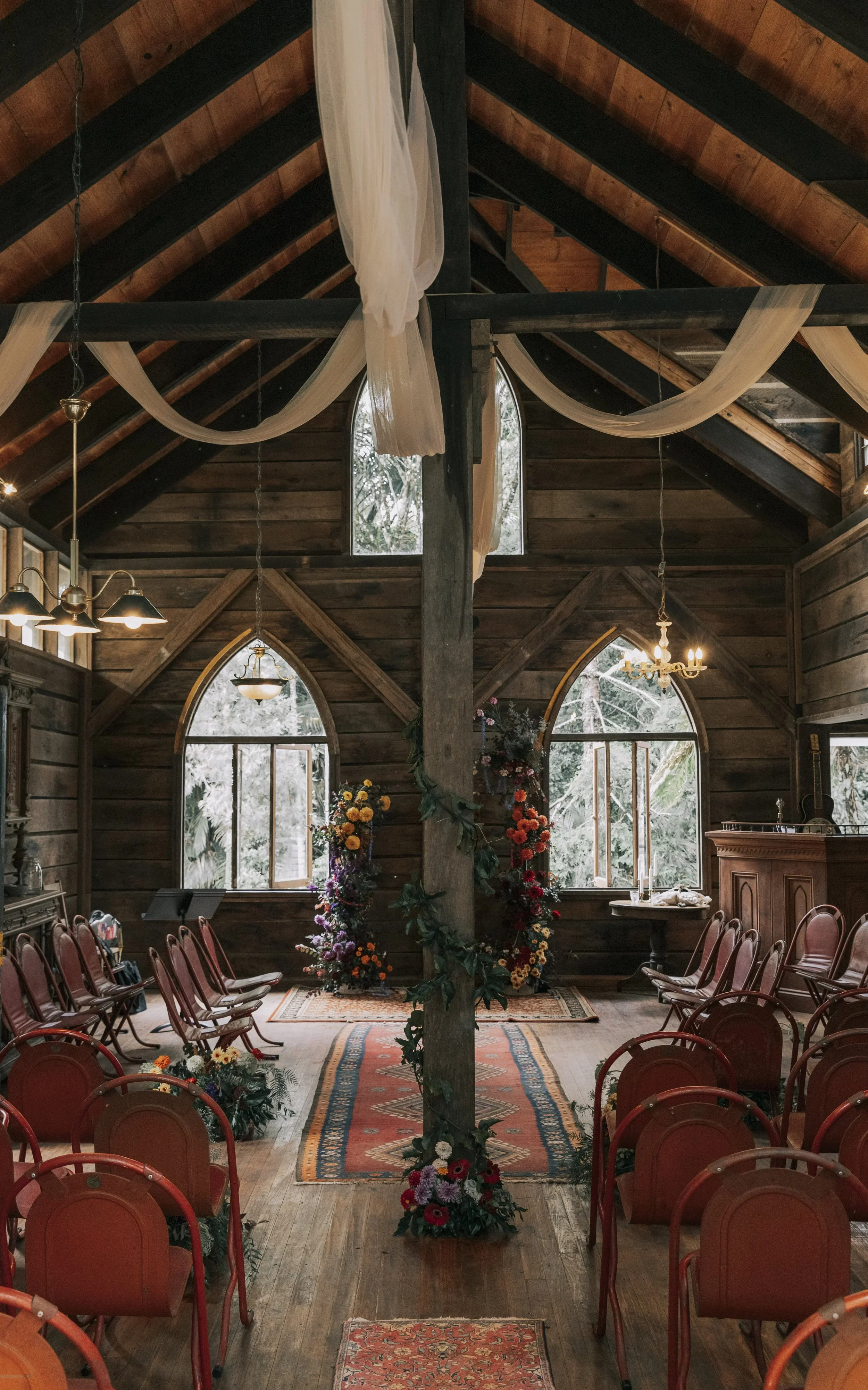 Interior of a rustic wooden church decorated for a wedding, with chairs arranged on either side of an aisle, floral displays near the windows, and fabric draping hanging from the ceiling beams.