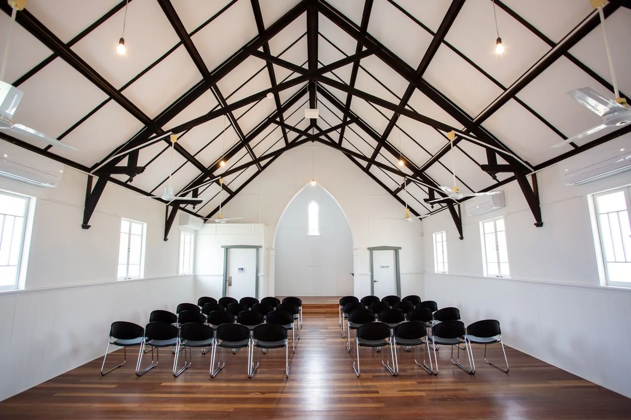 Empty indoor event space with wooden flooring, white walls, black chairs arranged in rows, high vaulted ceiling with exposed dark beams, multiple windows, ceiling fans, and hanging light fixtures.