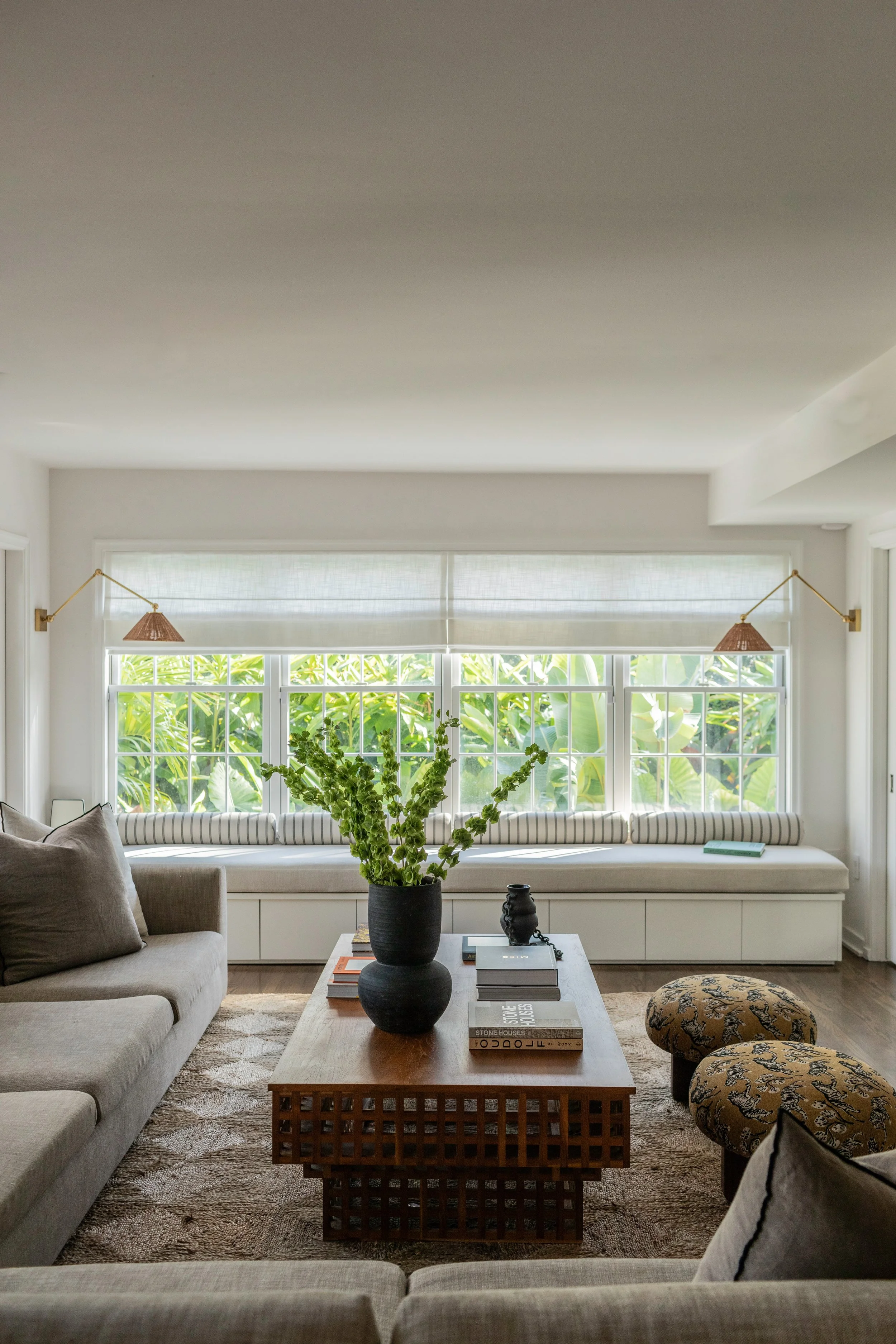 Living room with a beige sofa, a wooden coffee table with books and a black vase, two patterned ottomans, a built-in window seat with a cushion, and large windows with lush green plants outside.