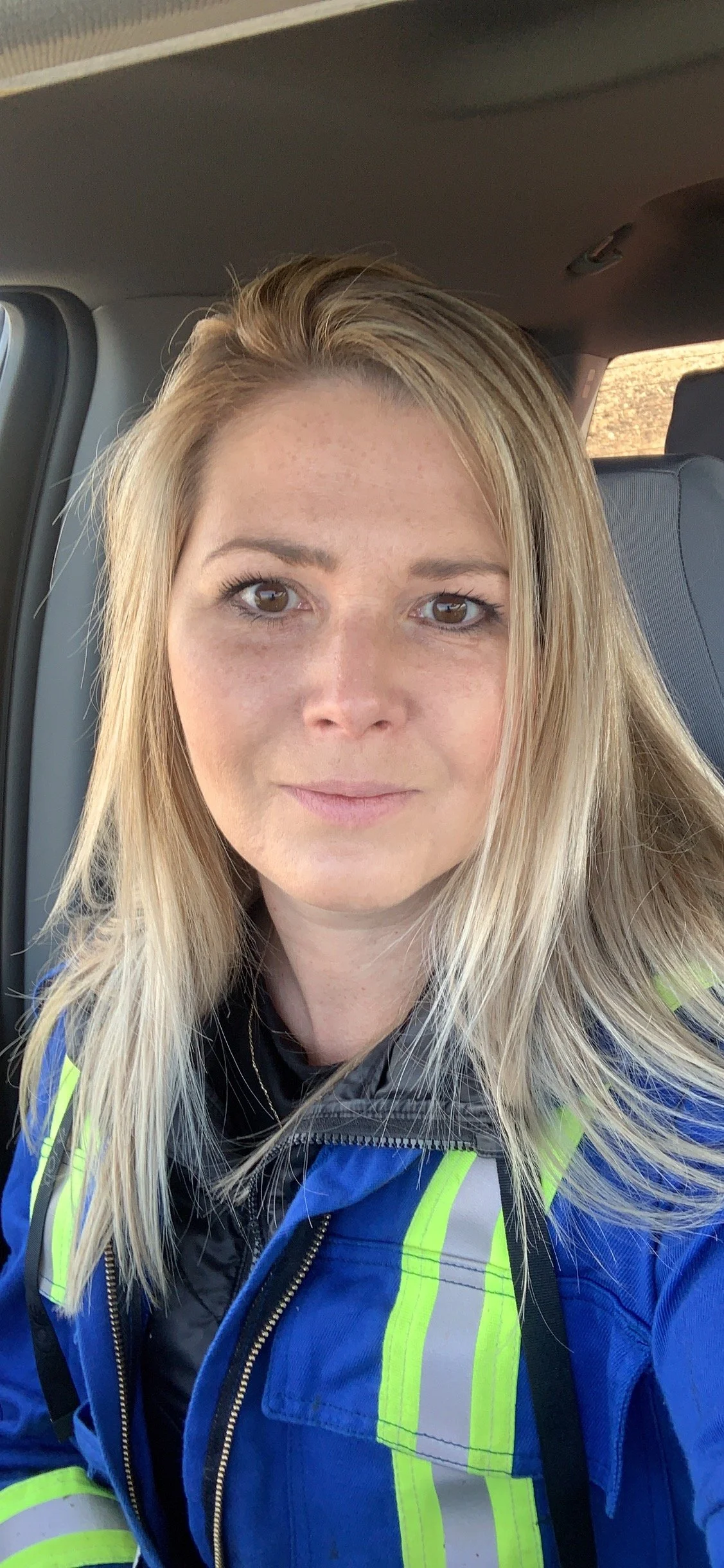 A woman with blonde hair and brown eyes, wearing a high-visibility blue jacket with yellow reflective stripes, sitting inside a vehicle.