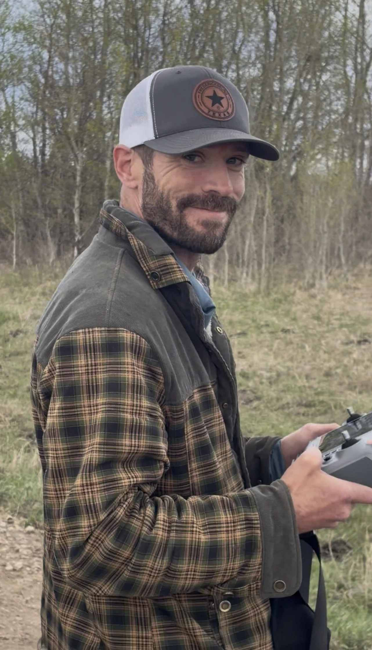 A man with a beard wearing a black and gray trucker hat with a star emblem, plaid shirt, and gray vest outdoors. He is holding a remote control and smiling, with trees and a partly cloudy sky in the background.