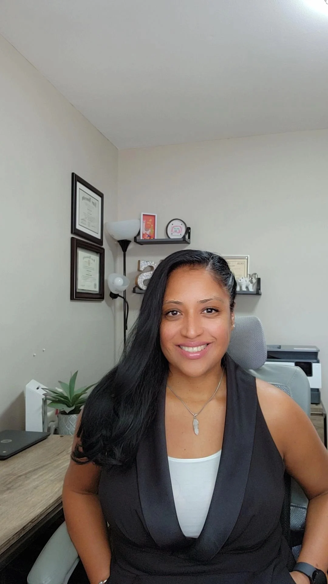 Therapist Shirley with long black hair sitting in her office chair in her home therapy office smiling.