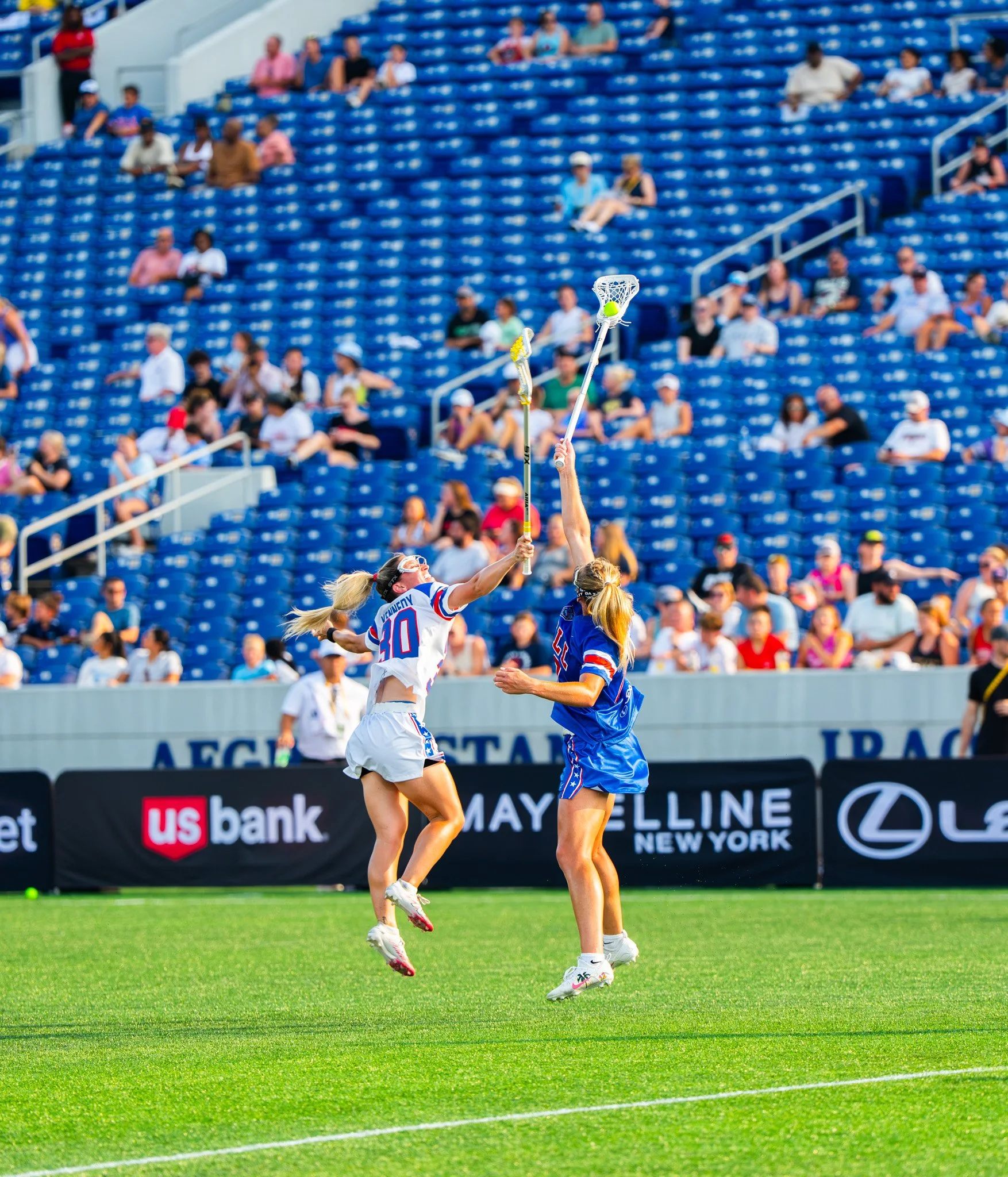 A lacrosse player wearing a white jersey and another wearing blue jump for a lacrosse ball in the women's lacrosse league. 