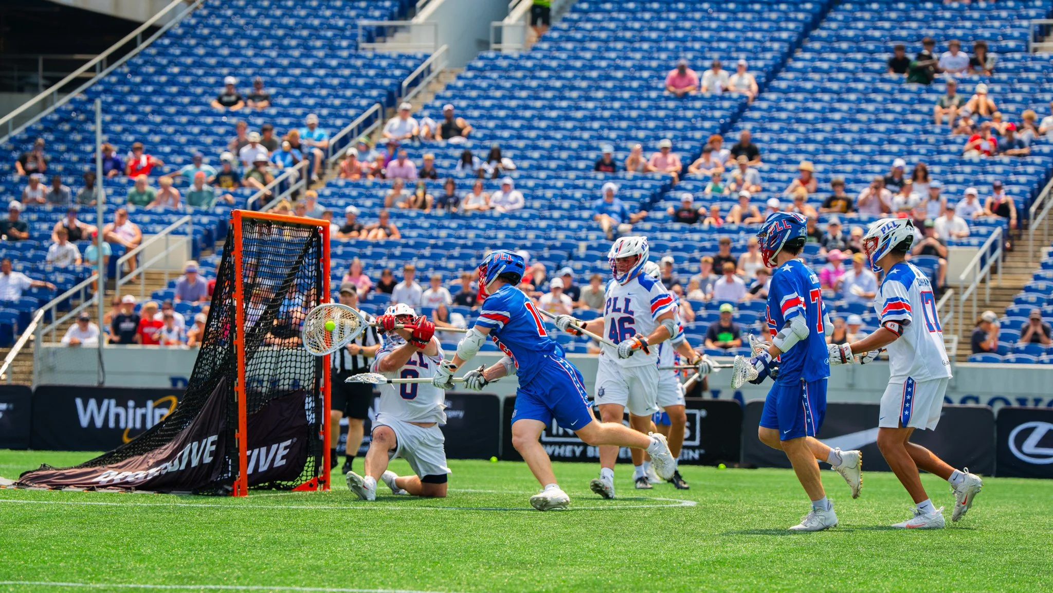 A lacrosse goalkeeper wearing a white jersey blocks a shot from a player wearing a blue jersey as players run around the goal in the professional lacrosse league. 