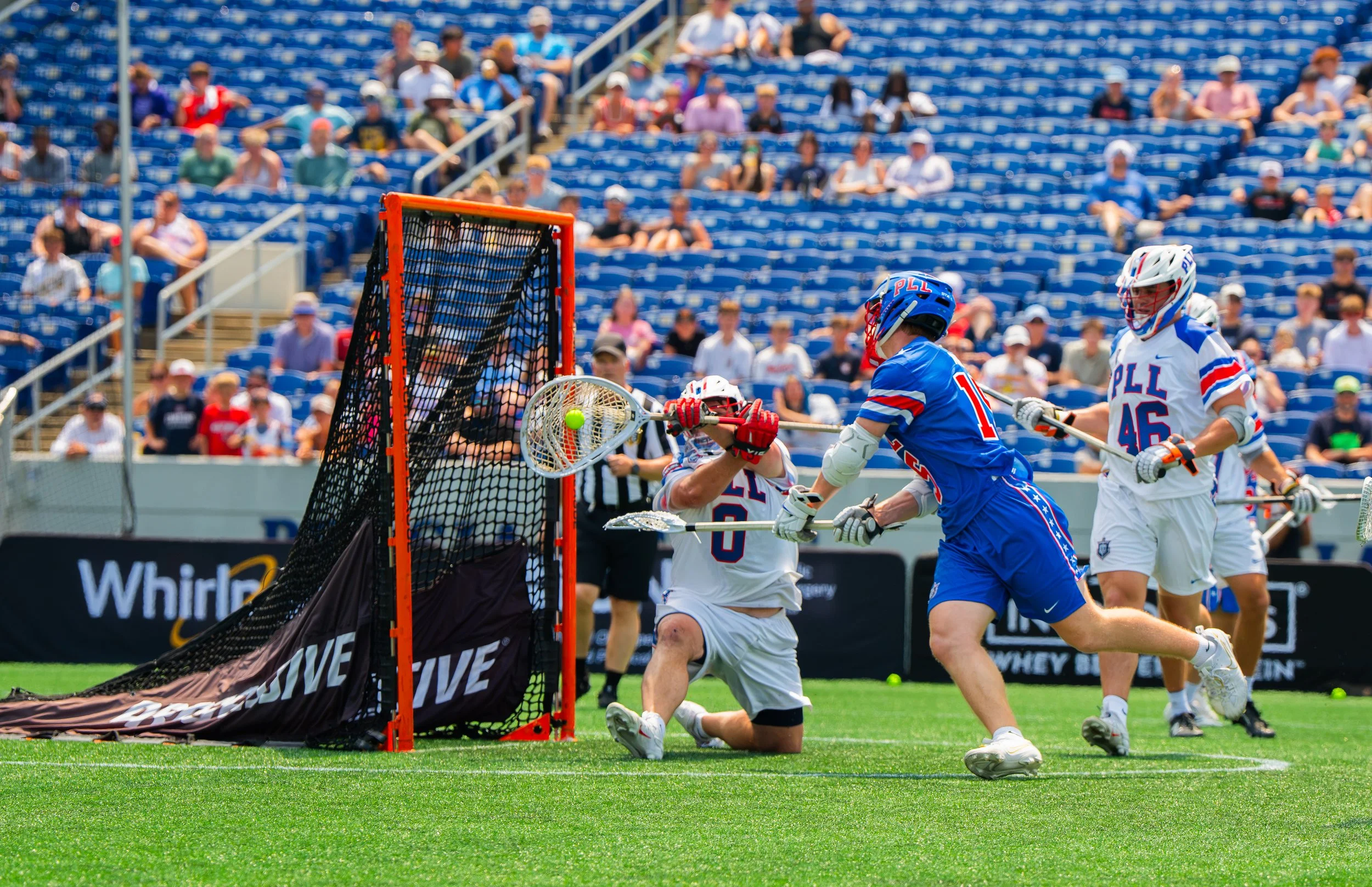 A lacrosse goalkeeper with a white jersey blocks the shot of a player wearing a blue jersey as other players watch on. 