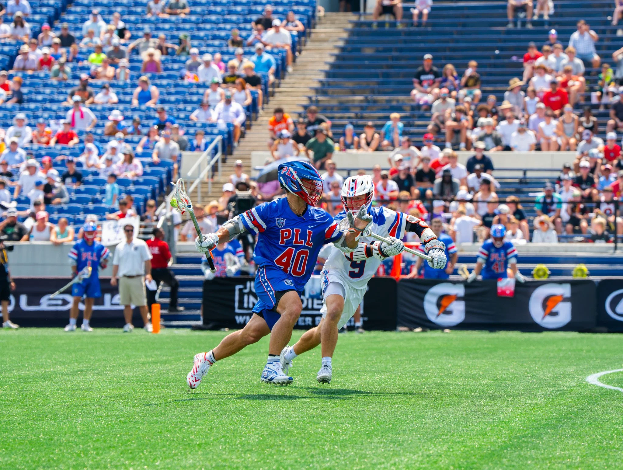 A lacrosse player wearing a white jersey runs alongside a player wearing a blue jersey in the professional lacrosse league. 