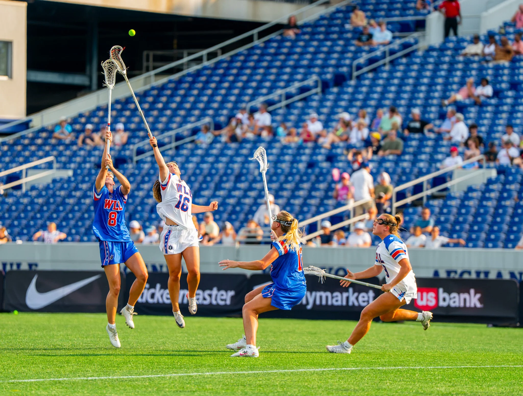 A lacrosse player wearing a white jersey and a player wearing a blue jersey reach their sticks into the air to catch a ball as two more players run near them in the women's lacrosse league. 