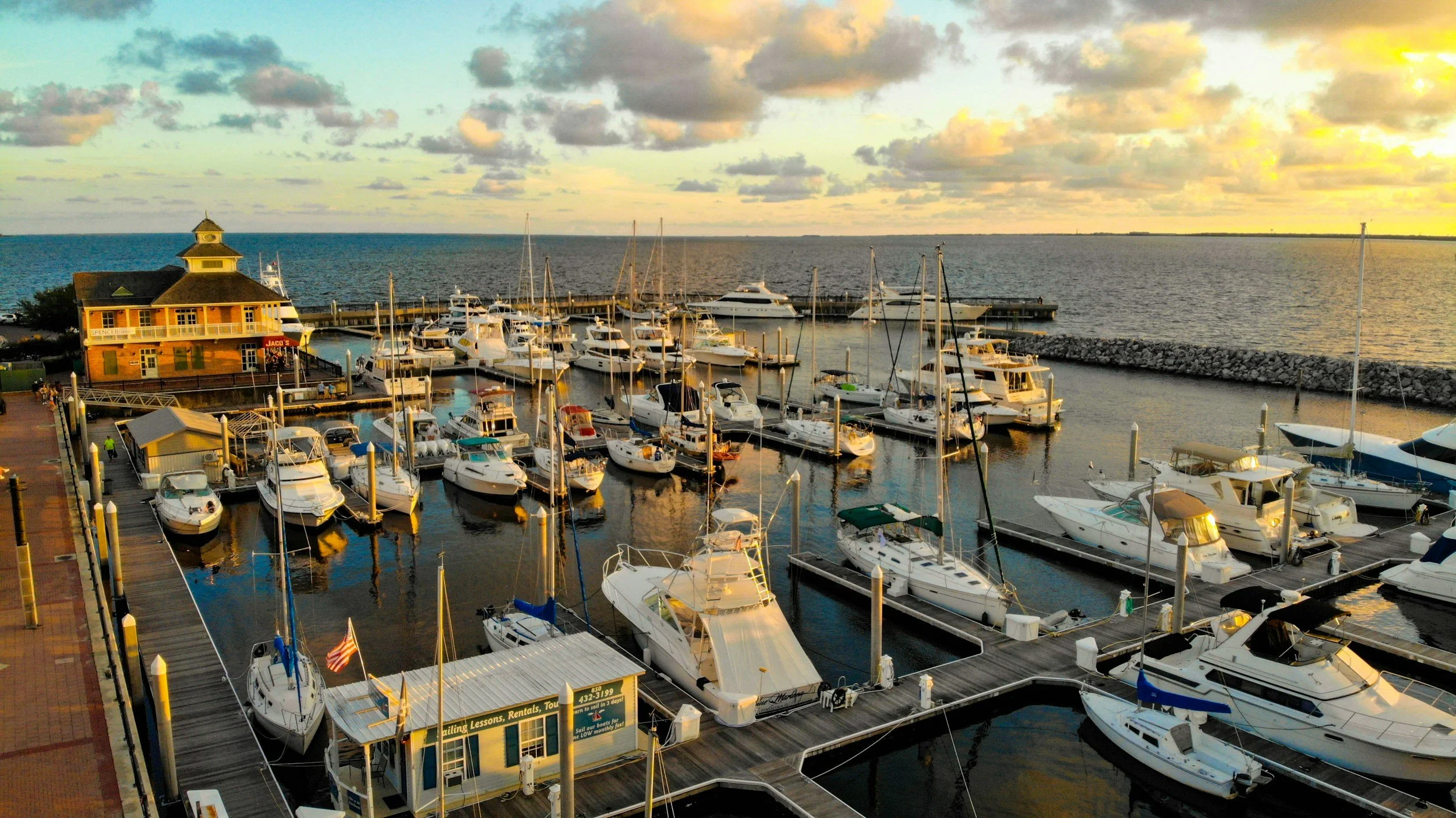 A marina filled with various boats and yachts docked at the piers at sunset. A building with a restaurant or shop is visible on the left, and the sky shows clouds with warm golden light from the setting sun over the water.