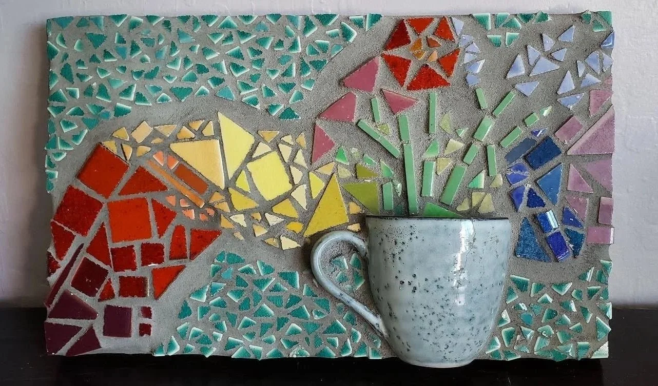 Colorful mosaic artwork featuring a white ceramic mug with green and yellow mosaic pieces appearing to rise from it, set against a multicolored background of small mosaic tiles in various shapes and colors.