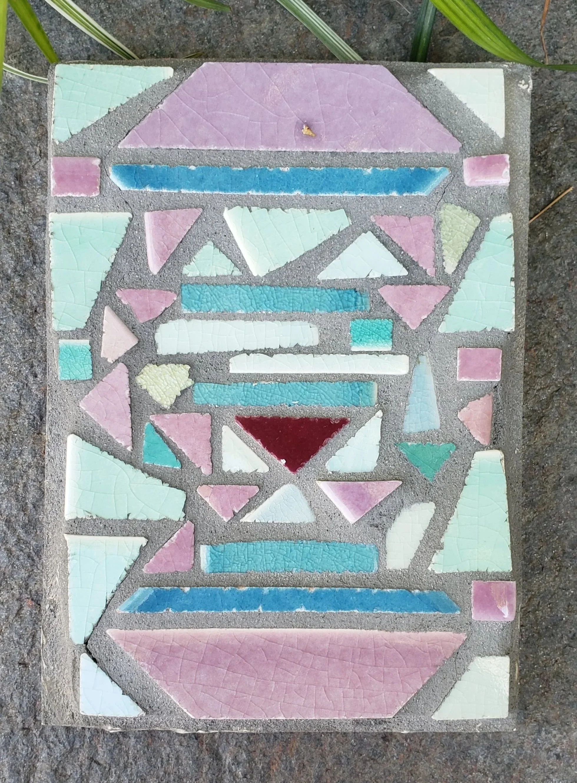 Colorful mosaic tile sidewalk art with geometric shapes and pastel colors on concrete surface surrounded by green plants.