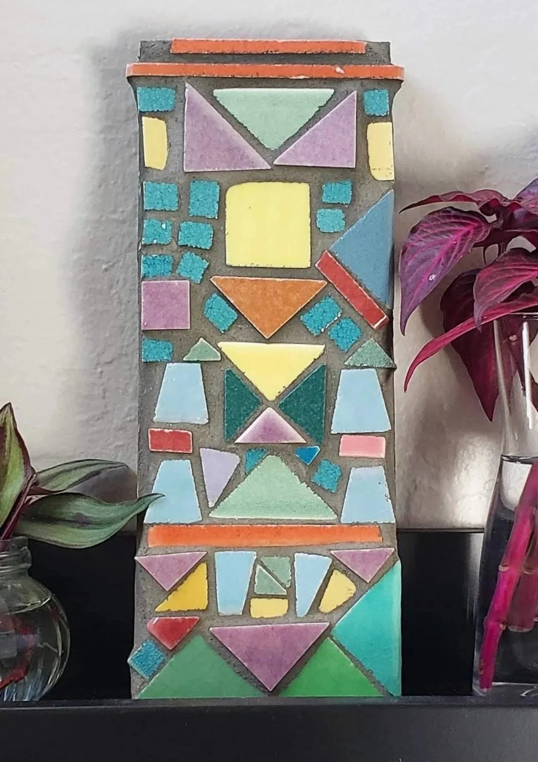 Colorful mosaic artwork with various geometric shapes, including squares, triangles, and rectangles, arranged vertically on a wall surrounded by plants.