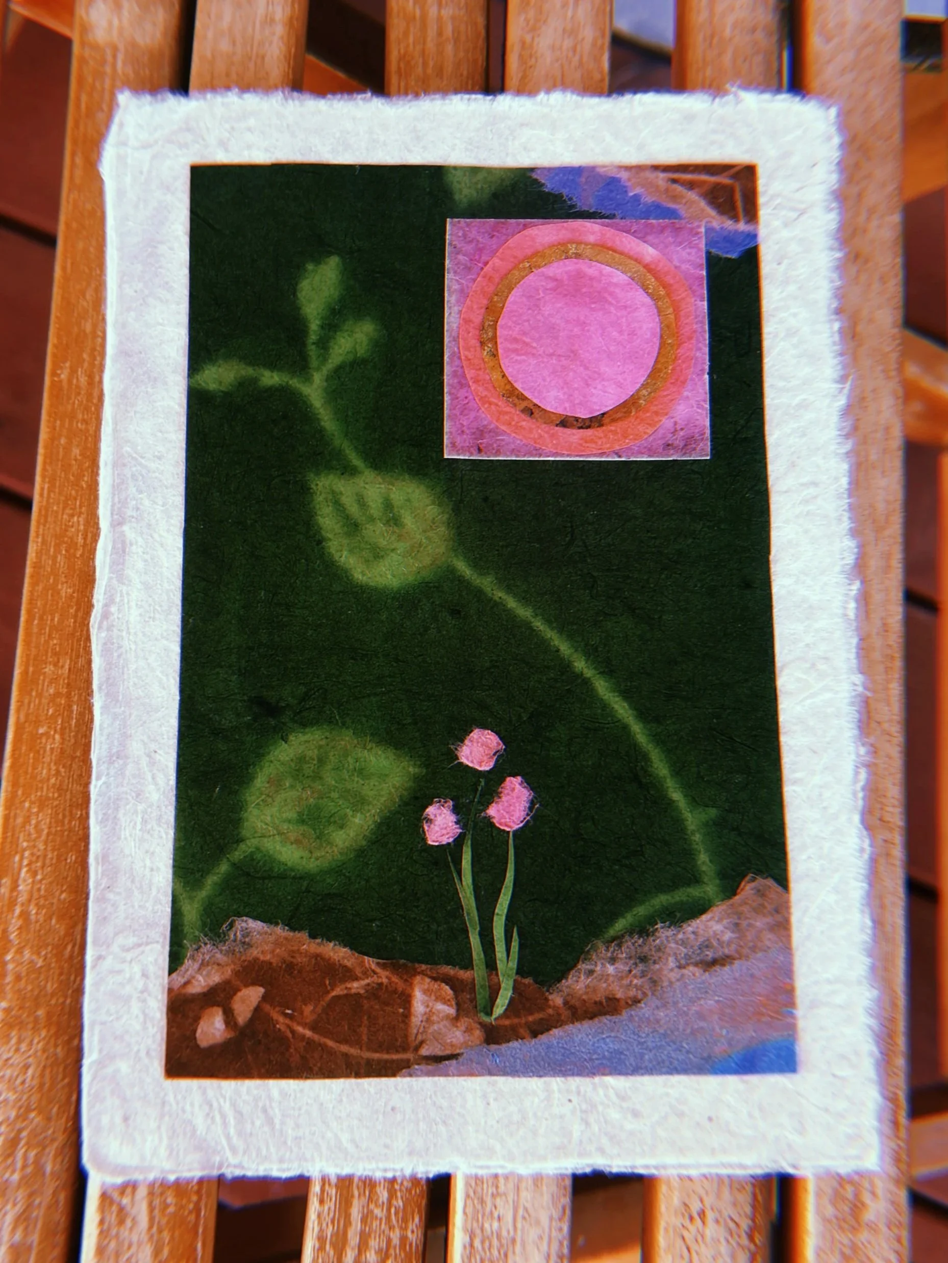 A handmade collage featuring a green leaf with three pink flowers and a pink circle with an orange border on a textured paper background.