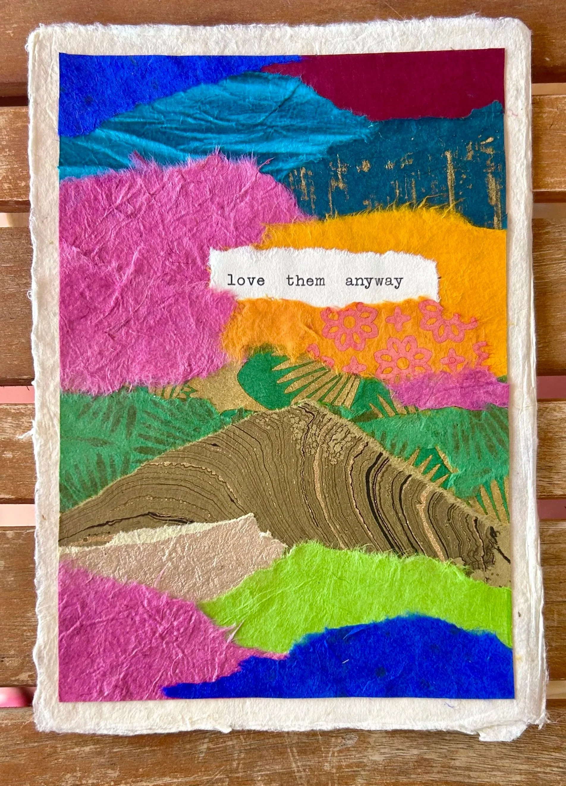 A colorful collage made of torn paper with mountains and trees in pink, blue, green, yellow, orange, brown, and gold, featuring a torn white paper with the words 'love them anyway' printed on it.