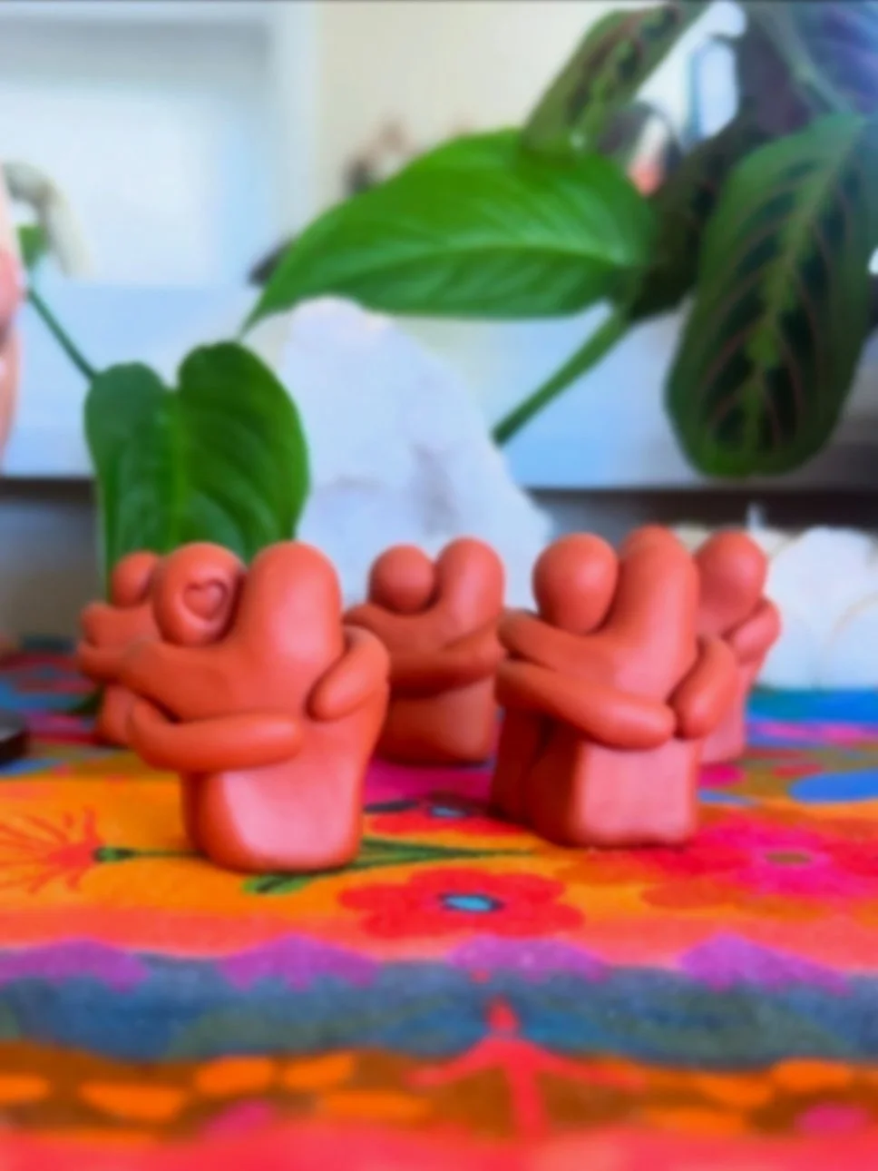 Colorful tablecloth with pink and orange shades, decorated with red, pink, purple, and blue patterns. Several pink ceramic hands in front, each with a finger pointing upwards or crossed, acting as a finger heart gesture. Behind the hands, there are t