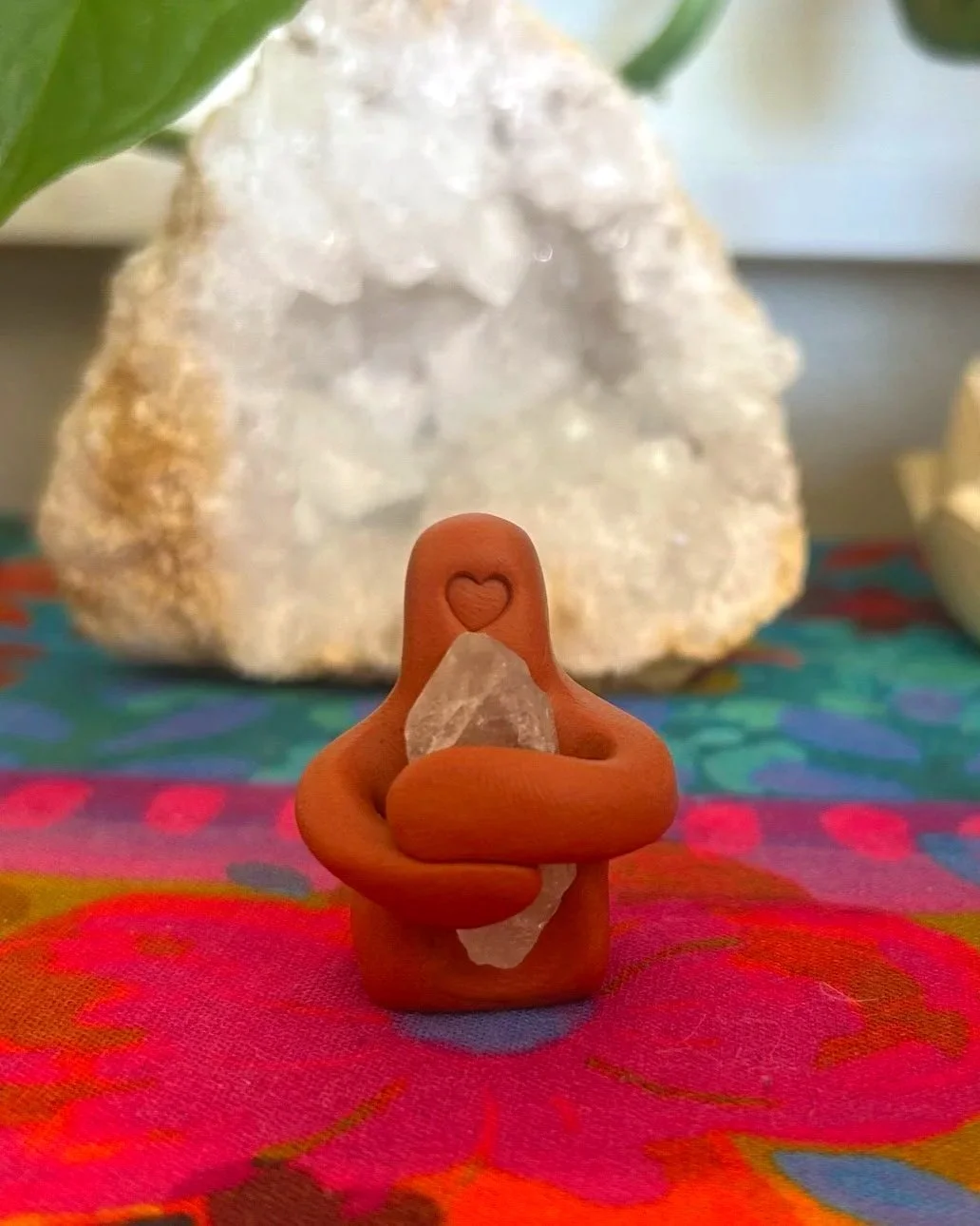 Small orange figurine with a heart engraved on its forehead, sitting cross-legged and holding a clear stone, placed on colorful fabric with a large mineral or crystal in the background.
