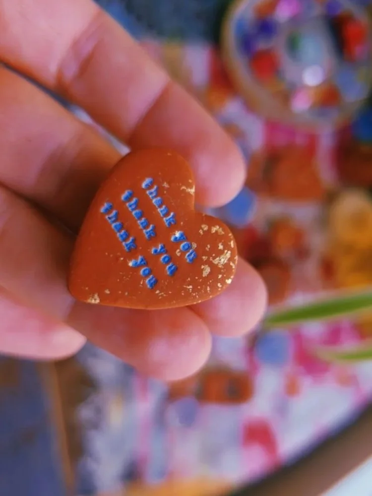 Close-up of a heart-shaped candy with blue text reading "For your pop heart" being held between fingers, with a blurred display of other candies in the background.