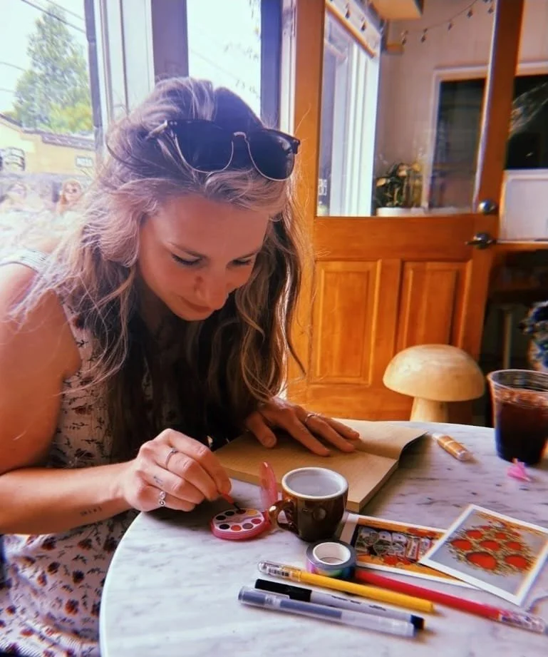 Woman with sunglasses on her head writing or drawing at a table with various pens, markers, and a journal, near a window in a cozy wooden interior.