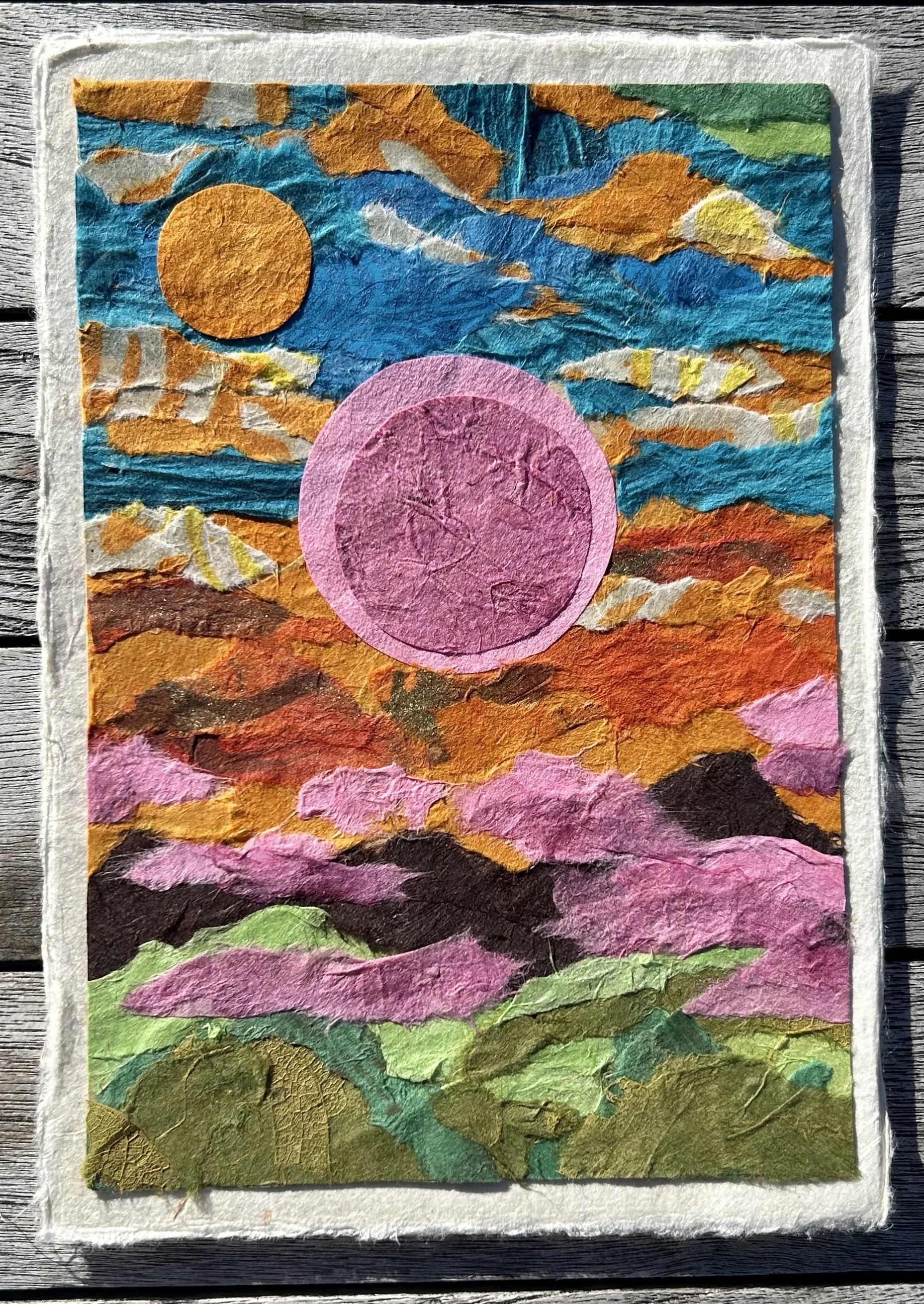A colorful collage of torn paper creating a landscape scene with a sky, mountains, and a prominent pink circle and yellow-orange sun.