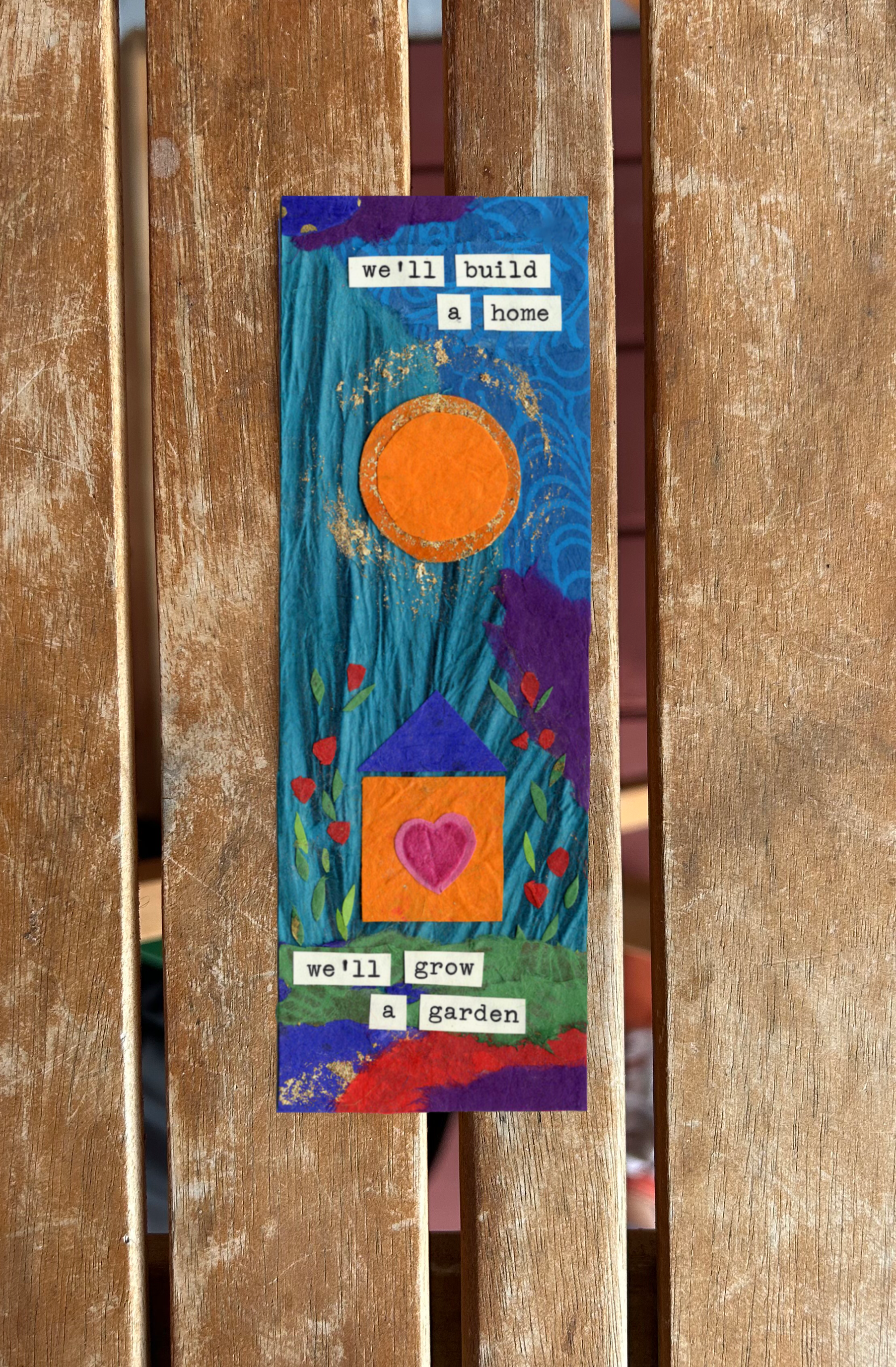 A colorful handmade fabric bookmark with a painted scene of a house with a purple roof, an orange wall with a pink heart, green grass, and abstract blue sky and orange sun. The bookmark has words that say "We'll build a home" at the top and "we'll gr