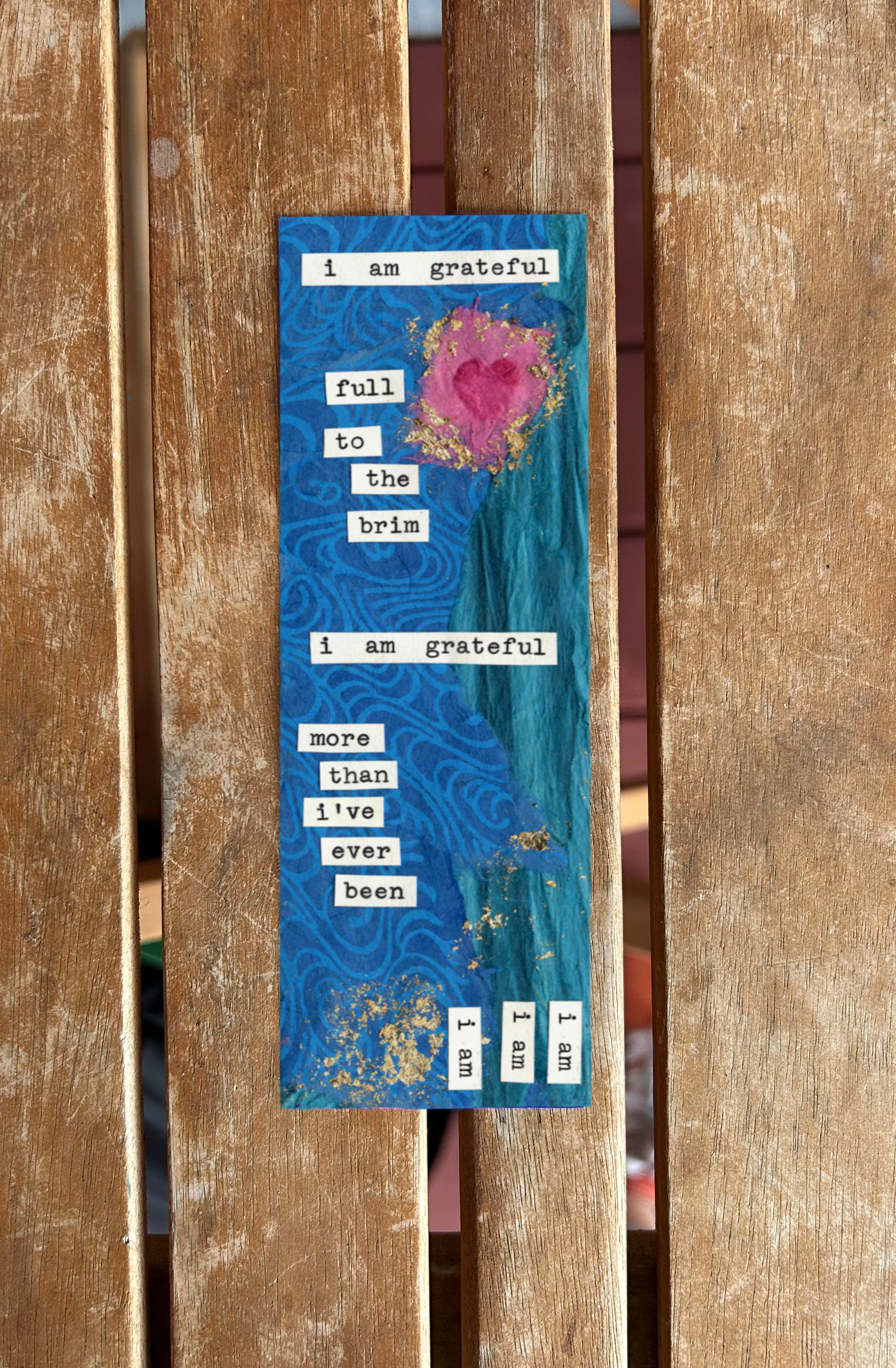 A colorful artistic bookmark with a blue background, gold splashes, and a pink and red heart shape at the top. The bookmark features cut-out words forming a sentiment about gratitude.
