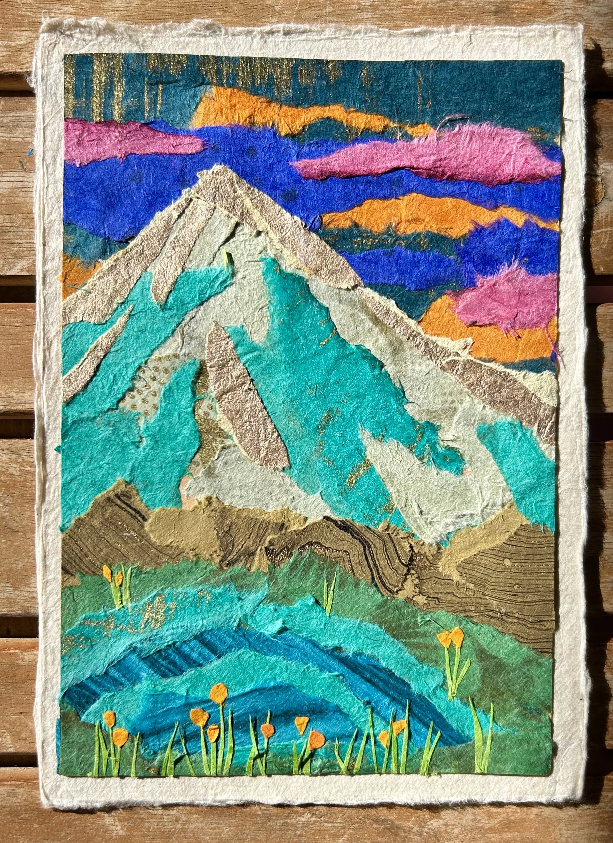 Colorful collage of a mountain landscape with a prominent snow-capped peak, vibrant sky with pink, orange, purple, and blue clouds, green rolling hills in the foreground, and flowers on the ground.